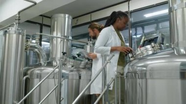 Çoklu etnik çeşitlilikte bira fabrikası çalışanlarının büyük çelik tanklarla birlikte çalıştığı, ekipmanların ayarlandığı ve bira yapma sürecini tartıştığı tam donanımlı çekim.