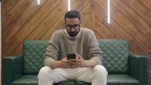 コワーキングオフィスでソファーに座って スマートフォンにメッセージを入力し 頭を回して遠ざけている若い思慮深い中東のビジネスマンの中間ショット — ストック動画