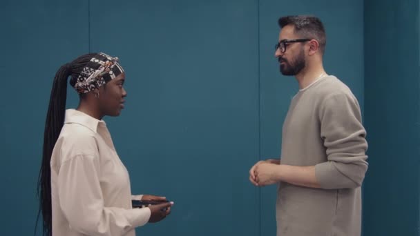 オフィスの青い壁の前に立っているカジュアルな服でアラブ人男性とアフリカ系アメリカ人女性のウエストアップショット お互いを見て笑顔と話す — ストック動画
