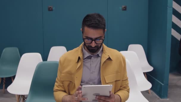 コワーキングオフィス施設の講演ホールに座って タブレットコンピュータを使用して メガネ カジュアルな黄色のジャケット シャツで忙しい中東の若い男のミディアムショット — ストック動画