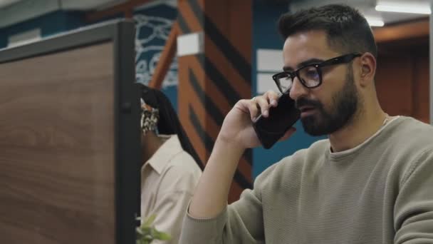 中特写照片 年轻的留着胡子的阿拉伯男子 戴着眼镜 坐在同事办公室的小隔间里 用智能手机和微笑交谈 以及背景中的黑人女性顾客 — 图库视频影像