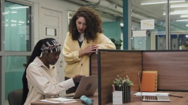 Orta ölçekli bir Afrikalı Amerikalı kadın stajyerin yeni işyerindeki masasında oturup laptopta çalışırken ve yanında duran Kafkas akıl hocası görevlerini açıklarken ve tavsiyelerde bulunurken.