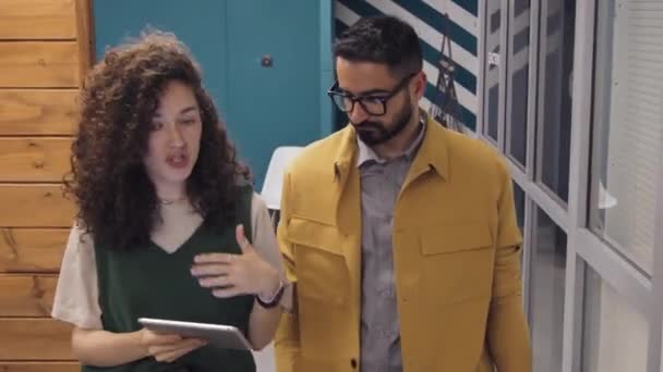 カーリーヘアの若い白人女性のミディアム追跡ショット タブレットコンピュータを保持し コワーキングオフィス施設を歩き 作業計画を議論中 眼鏡で中東男性をひげました — ストック動画