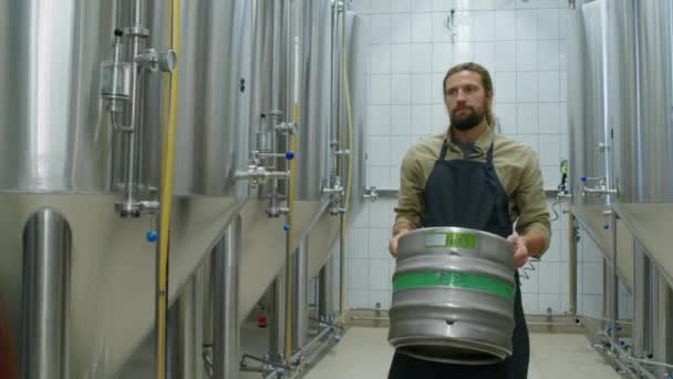 赤ひげを持つ若い白人男性のミディアム追跡ショット エプロンでゆっくり発酵タンクを歩き ゲージを見て 新鮮なビールの重いケグを運ぶ — ストック動画