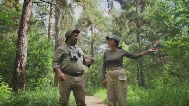 多民族の男性森林レンジャーの中型ショット カーキのユニフォーム 双眼鏡とウォルキートーキー 自然公園に立ってヒスパニックの女性同僚とチャット — ストック動画