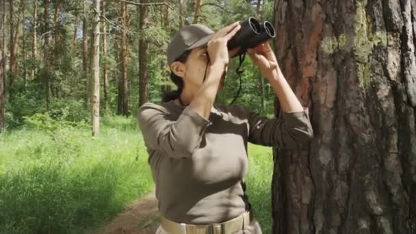 森の松の隣に立っている若いヒスパニック系女性生物学者の中型肖像画 双眼鏡を通して鳥や動物を見ながら 静かな笑顔でカメラを見る — ストック動画