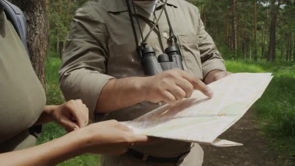 カーキのユニフォームで認識できない男性の森の警備員の手と体のクローズアップショット 自然公園で匿名の女性のハイカーを助け 地図上のルートを指し 方向を説明する双眼鏡 — ストック動画