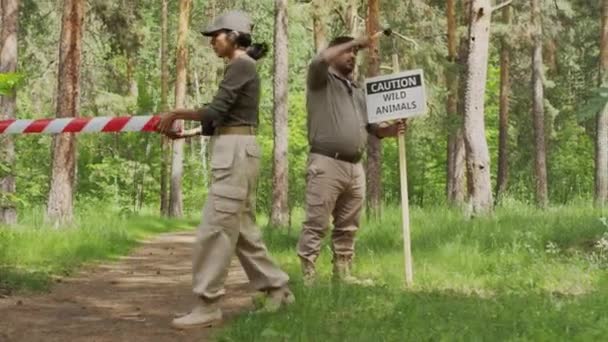 カーキのスウェットシャツとユーティリティパンツでヒスパニックの女性の木材のフルショットは バリケードテープで森林地帯を切り離し インドの男性同僚は野生動物の警告サインを掲げています — ストック動画