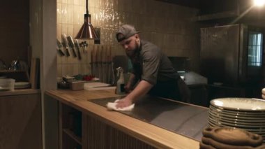 Genç, sakallı, beyaz erkek pizza şefi. Ceket, önlük ve şapka giymiş. Loş aydınlatılmış bir lokanta mutfağında vardiyaya başlıyor.