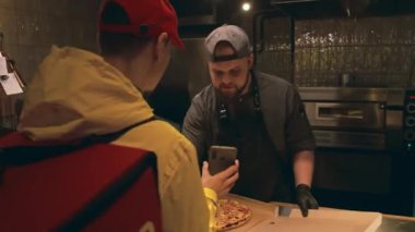 Sırt çantasıyla restorana giren yemek servisi kuryesinin orta boy fotoğrafı. Karton kutuda pizza paketleyen ve hem gülümseyen hem de sohbet eden erkek şefe siparişini gösteriyor.