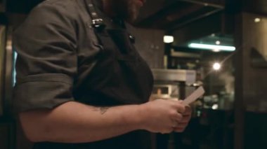 Orta boy yakın plan çekimler, önlüklü ve şapkalı beyaz erkek şefin restoran mutfağında sipariş kâğıdı alıp, okuduktan sonra taze sebzeler hazırlamaya başlaması.