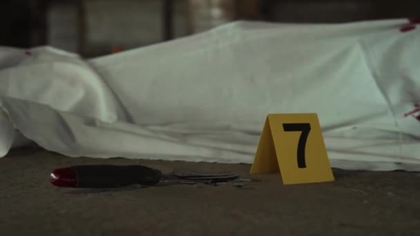 殺人現場で白いシートで覆われた死体のクローズアップショット その横に血が横たわるキッチンナイフ そして黄色いフォレンジックマーカー7 — ストック動画
