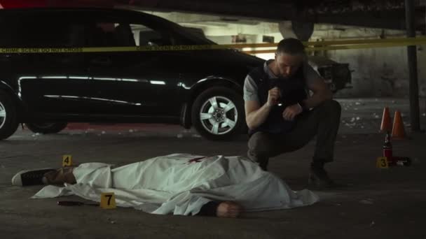 白いシートで覆われた死体を検査し 暗い地下駐車場で殺人現場を調査する 防弾ベストとトーチを保持する白人の男性警察探偵のフルショット — ストック動画