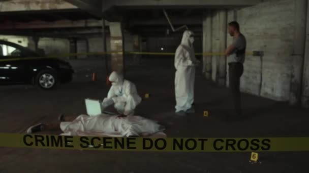 在城市停车场的凶杀现场工作的身着防护服的警察侦探和法医调查员的全照 尸体上铺着白纸 前面还挂着黄色的隔离带 — 图库视频影像