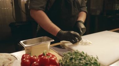 Restoran mutfağında çalışan şef üniformalı, şapkalı ve eldivenli meşgul beyaz bir adamın orta ölçekli yüz ve elleri ve kesilmiş peynirli mozzarella topunu doğrayan tahta üzerinde bıçakla.