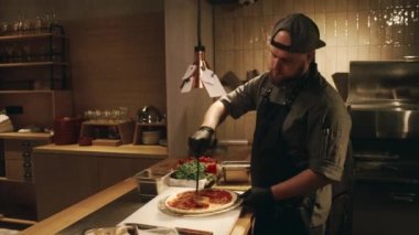 Üniformalı, önlüklü, şapkalı ve eldivenli beyaz bir erkek aşçının pizza tabanına domates sosu sürerken, dairesel hareketlerle etrafa yayılışını ve üstüne bir avuç rendelenmiş peynirle...
