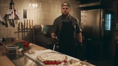 Kafkasyalı erkek aşçının restoran mutfağında çalışırken, uzun saplı pizza kabuğunu alıp, tezgahtan tepsiyi alıp, fırını açarken ve yemek pişirmek için içeri yerleştirirken.