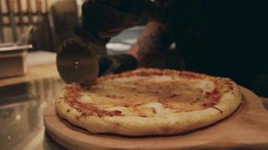 Restoranın mutfağında, ahşap tabakta duran yeni pişmiş peynirli pizzanın yakın plan çekimi ve onu dilimleyen isimsiz bir şefin elleri.