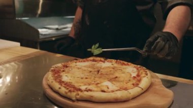 Yakından çekilmiş taze pişmiş ve dilimlenmiş peynirli pizza gurme restoranının mutfağında tezgahta duruyordu ve tanınmayan siyah eldivenli erkek aşçı ortaya fesleğen yaprağı koyuyordu.