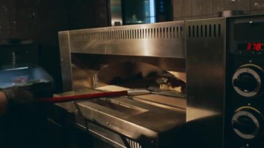 Siyah üniformalı ve eldivenli tanınmayan şefin, fırından yeni çıkmış yıldız şekilli pizzayı metal kürekle alıp atölyenin tepesine koyduğu orta boy bir fotoğraf.