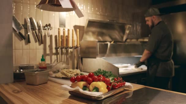 新鮮なチェリートマト ピーマン ハーブがレストランのキッチンのワークデスクに横たわる金属トレイの完全な選択的なフォーカスショット および男性シェフはぼやけた背景でピザ生地をポンド — ストック動画
