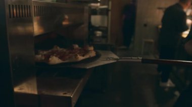Erkek şefin fırından yeni çıkmış, dumanı üstünde, yıldız şeklinde pizza yerken, pizza küreklerini kullanırken, loş ışıklandırılmış restoran mutfağında ve arka planda çalışan diğer meslektaşlarının yakın plan fotoğrafları.