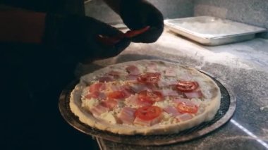 Siyah önlüklü ve eldivenli isimsiz bir şefin jambonun üstüne dilimlenmiş domates ve rendelenmiş peyniri sererken restoran mutfağında pizza yaparken yakın plan çekimleri.