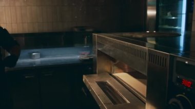Tanımlanamayan erkek şefin, pizza kabuğu kullanarak, loş aydınlatılmış restoran mutfağında, profesyonel fırına pizza yerleştirirken yakın plan çekimleri.