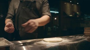 Şefin üniformalı, siyah önlüklü, tanınmayan bir adamın el ve gövdesinin orta boy yakın çekimi konteynırdan bir avuç un alıp atölyedeki pizza hamuruna serpiyor.