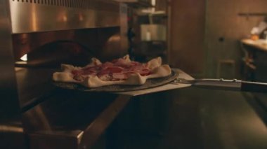 Ekran dışı şefin yakın plan görüntüsü yıldız şeklindeki pizzayı pişirmek için taş levhanın üzerine koyuyor, sonra da metal kürek kullanarak kapıyı kapatıyor.
