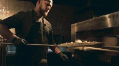 Siyah üniformalı, önlüklü ve eldivenli beyaz erkek şefin metal kürekte pişmemiş pizzayı pişirmek ve kapamak için sıcak fırına koyduğu orta boy fotoğrafı.