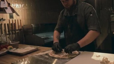 Kafkasyalı siyah üniformalı, eldivenli, restoran mutfağında pizza yapan ve pizza tabanında rendelenmiş peynirin üzerine dilimlenmiş mantar seren beyaz aşçının yakın plan fotoğrafını çek.
