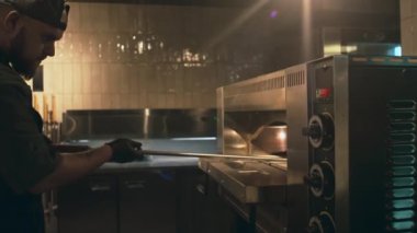 Genç aşçının metal kürekle fırına uzandığı, buharı tüten sıcak pizzayı çıkardığı, onu uzattığı ve kameraya gösterdiği orta boy bir fotoğraf.