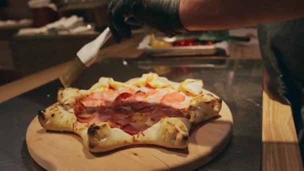 ブラックグローブでプロの男性シェフの手のクローズアップショット 溶けたバターを広げ スペシャルティピザのトーストクラストにブラシをかけ キッチンのカウンターに木製の板に横たわる — ストック動画