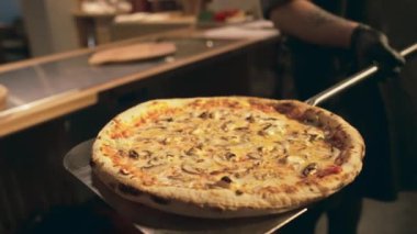 Lokanta mutfağında taze pişmiş mantar ve peynirli pizzanın yakın plan çekimi ve kamera için elinde tutan isimsiz bir şefin elleri.