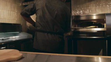 Siyah üniformalı, önlüklü ve eldivenli, fırından yeni çıkmış peynirli pizzayı metal kürekle alıp tezgahtaki tahta tepsiye koyan tanınmamış bir aşçının orta boy fotoğrafı.