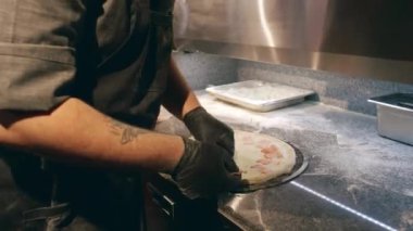 Kolunda siyah üniforma ve eldivenlerle restoranda pizza yaparken rendelenmiş peynirin üzerine dilimlenmiş domuz pastırması seren beyaz bir aşçının orta boy yakın çekimi.