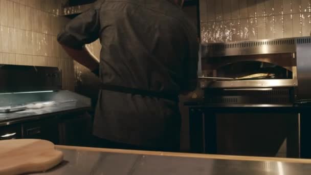 身着黑色制服 围裙和手套的大厨用铁锹从烤箱里取出新鲜烤的奶酪披萨 放在柜台上的木制盘子里 拍了中镜头 — 图库视频影像