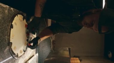 Kafkasyalı siyah üniformalı ve önlüklü bir aşçının loş bir restoranın mutfağında yıldız şeklinde pizza yaparken orta boy dikey pozu ve pizza tabanında vişneli domates sererken.