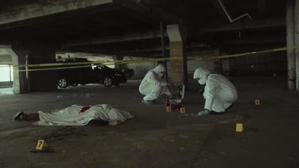 都市駐車場の殺人現場でシートで覆われた人間の死体のフルショット 2人の法医学捜査官が証拠を収集し コンクリートの床にサンプルを集め 警察のライトを点滅させた — ストック動画