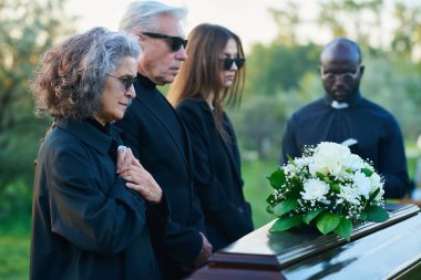 Cenaze töreni sırasında kapalı kapaklı tabutun önünde ellerini göğsünde tutan olgun bir kadının yasını tutuyordu.