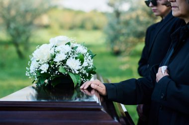 Cropped shot of mature woman and her family in mourning attire standing in front of coffin with closed lid with fresh white chrysanthemums clipart