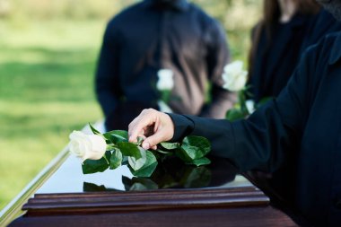 Hand of mature woman in mourning attire putting white rose on top of closed coffin lid while standing in front of camera against other people clipart