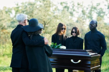 Tabutun önünde dikilen siyah elbiseli olgun yas tutan çifte ve cenazede yas tutan bir grup kültürlü insana odaklan.