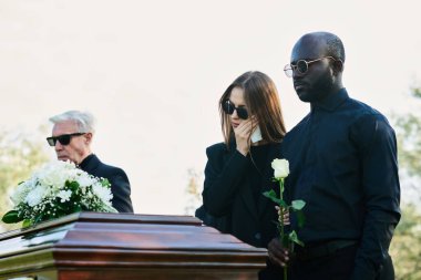 Beyaz güllü genç adam, aile üyesinin cenazesinde siyah elbiseli ve güneş gözlüklü karısının yanında gözyaşlarını mendille siliyor.