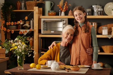 Mutlu kıdemli kadın örgü hırka giyip tatlı, gülen torununu örgü örgülü saçlarıyla kucaklarken masada çay ve turta ile oturuyor.