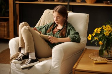 Gündelik kıyafet giymiş, büyük annenin evinde rahat bir koltukta otururken sayfalarca açık kitaba bakan tatlı bir kız öğrenci.