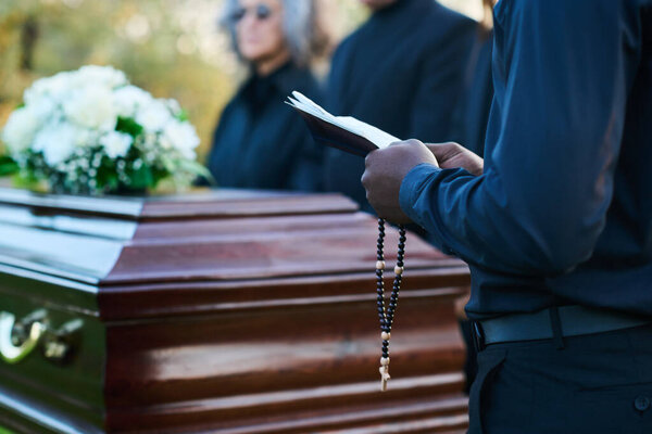 Close-up of young African American priest with open Holy Bible and rosary standing in front of coffin and relatives of passed away person