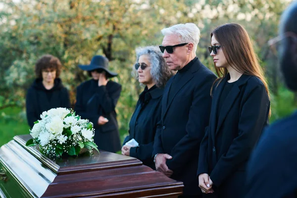 Family of three grieving people in sunglasses and mourning clothes standing in front of coffin with white flowers on lid during funeral service