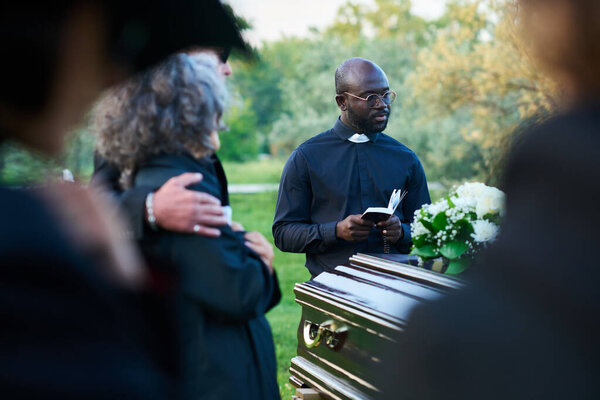 Focus on young pastor with Holy Bible and rosary beads standing in front of coffin with dead person and group of relatives during funeral service